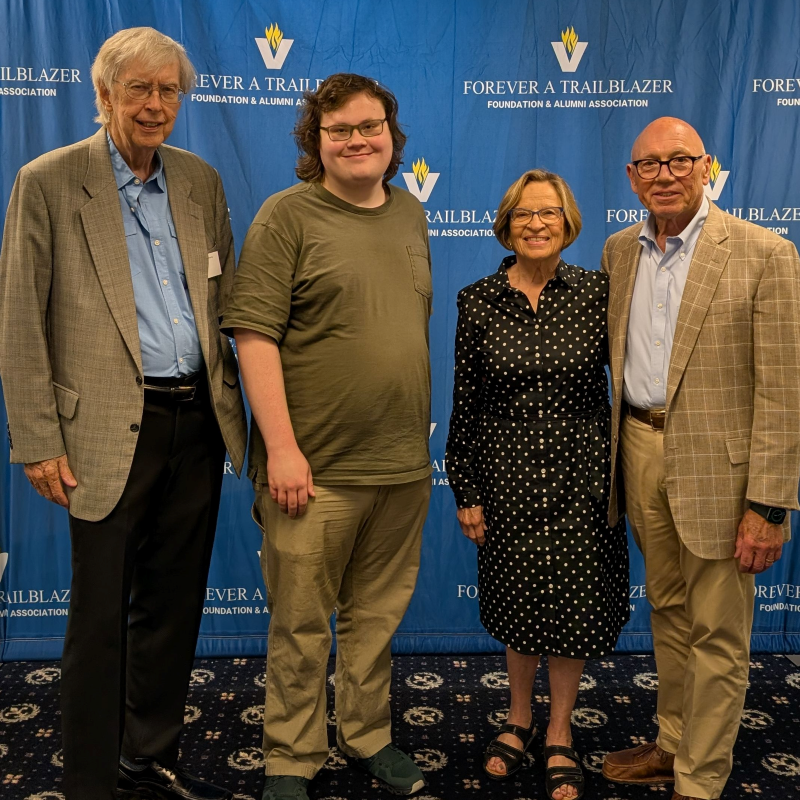 Four people pose together at a Forever a Trailblazer Foundation event, smiling before a blue logo backdrop.